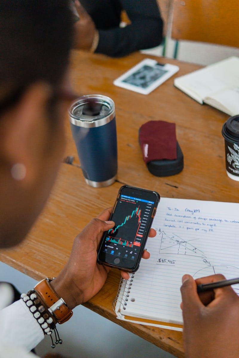 A person reviewing stock market data on a smartphone while taking notes during a meeting.